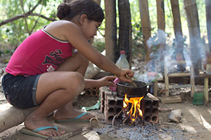Foto de uma menina brincando de cozinhar com um fogo acesso, em volta do fogo tem tijolos que seguram a panela de metal, a garota esta sentada em um galho de madeira.