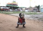 Foto com um garoto abaixado, vestindo uma máscara de caveira com dois chifres, segura um graveto com sua mão direita.