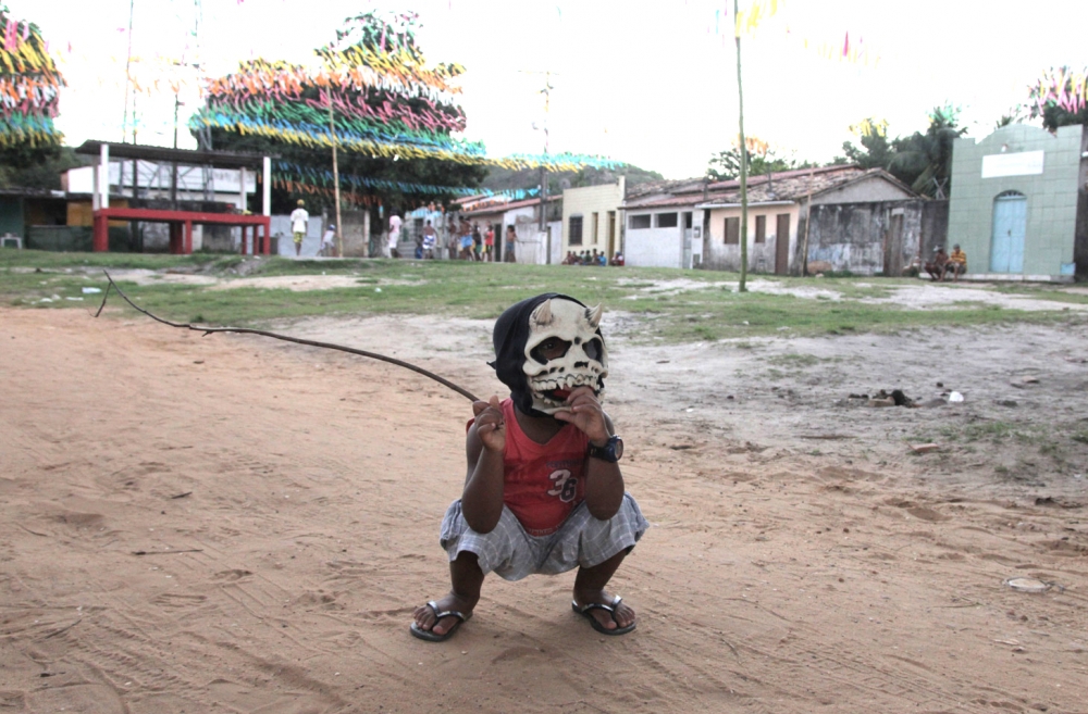 Foto com um garoto abaixado, vestindo uma máscara de caveira com dois chifres, segura um graveto com sua mão direita.