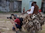 Foto de um homem tirando uma foto, de traz dele um homem negro fantasiado de senhor de escravos, aponta um rifle falso para a nuca do fotografo.