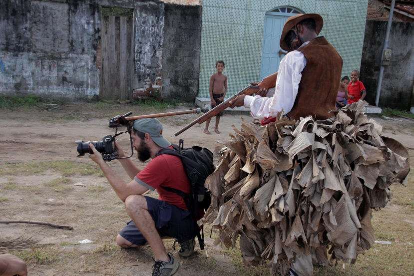 Foto de um homem tirando uma foto, de traz dele um homem negro fantasiado de senhor de escravos, aponta um rifle falso para a nuca do fotografo.