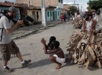 Foto de um homem filmando dois garotos negros, com a cara pintada de preto estão vestidos de escravos, com cordas amarradas sobre os pulsos eles fazem gestos de implorar, uma criança segura a corda dos dois meninos, ele está vestido como senhor, ele está com a cara pintada de preto.