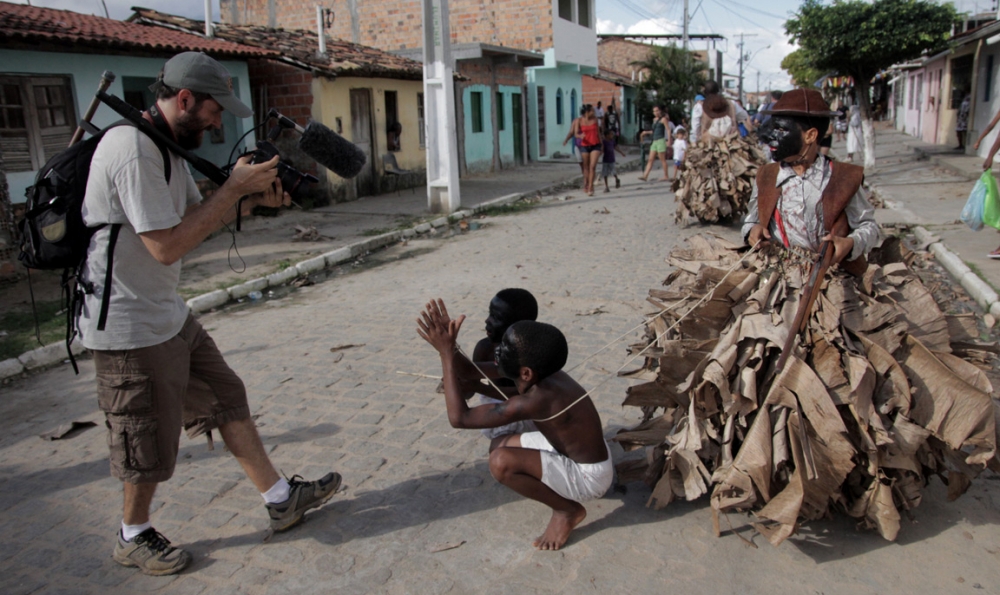 Foto de um homem filmando dois garotos negros, com a cara pintada de preto estão vestidos de escravos, com cordas amarradas sobre os pulsos eles fazem gestos de implorar, uma criança segura a corda dos dois meninos, ele está vestido como senhor, ele está com a cara pintada de preto.