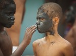 Foto composta por dois garotos, onde o primeiro garoto negro está com a cara cheia de lama, passa lama na cara do segundo garoto com uma expressão séria.