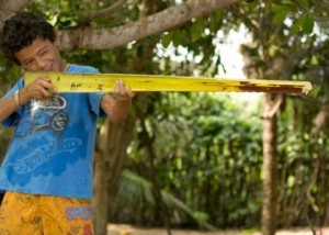Foto de um garoto segurando uma folha de um coqueiro cortada só com seu meio, o garoto está segurando como se fosse um rifle, com um olho aberto e outro fechado ele mira.