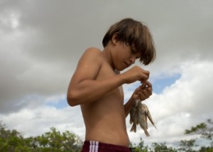 Foto de um garoto sem camisa, ele está segurando vário peixes em sua mão.