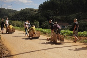 Foto ilustra várias crianças brincando com carrinhos de madeira, onde uma puxa o carrinho e outra fica em cima.