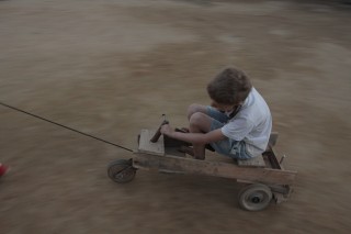 Foto com um garoto brincando em cima de um carrinho de madeira está sendo puxado por um cabo amarrado ao carrinho.