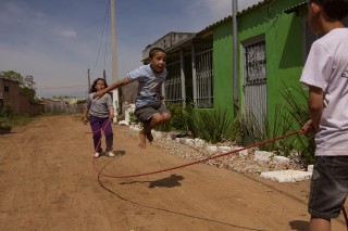 Foto demonstra três crianças brincando de pular corda, um garoto esta saltando, enquanto outras duas crianças, um menino e uma menina, estão batendo a corda.