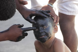 Foto de uma pessoa passando tinta no rosto todo de um garoto.