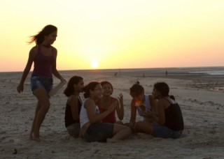 Foto ilustrando garotas sentadas em um circulo enquanto uma esta correndo em volta com um chinelo na mão ao por do sol.