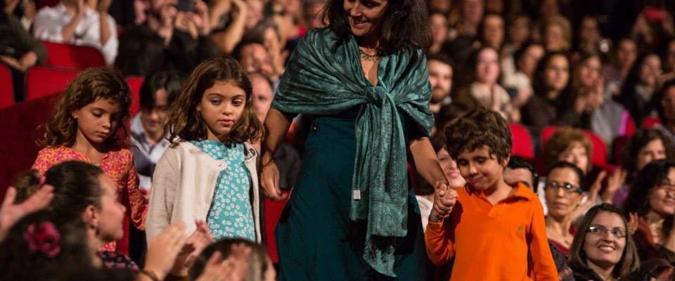 Foto de um salão cheio de pessoas sentadas observando uma mulher descer as escadas, de mãos dadas a uma menina e um menino.