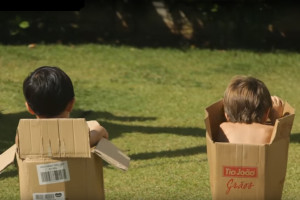 foto de duas crianças de costas, brincado dentro de caixas de papelão, onde o garoto da direita se inclina dentro de sua caixa.