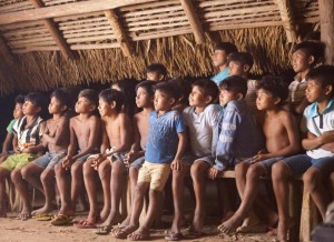Foto compostas de varias crianças sentadas e observando, dentro de uma casa feita de palha.