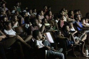 Foto de um salão cheio de crianças sentadas e observando um telão, todas as crianças estão rindo.