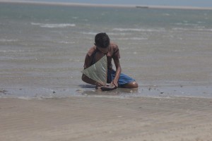 Foto de um garoto que a beira do mar, ajoelhado brinca com seu barco a vela de brinquedo.