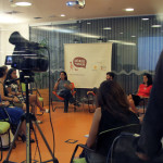 Foto composta por várias pessoas sentadas em círculo, ao fundo uma mulher fala ao microfone, uma câmera esta os filmando.