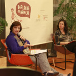 Foto com duas mulheres sentadas, uma senhora falando ao microfone, ao seu lado esquerdo uma mulher observa a senhora falar.