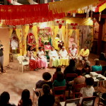 Foto ilustrando  o público total de uma palestra ,onde ao fundo há um palco composto de quatro mulheres e dois homens falando a um número de pessoas sentadas assistindo ao evento.