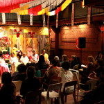 Foto ilustrando  o público total de uma palestra ,onde ao fundo há um palco composto de quatro mulheres e dois homens falando a um número de pessoas sentadas assistindo ao evento.