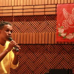 Foto onde há um homem negro com um microfone na mão falando ao público.